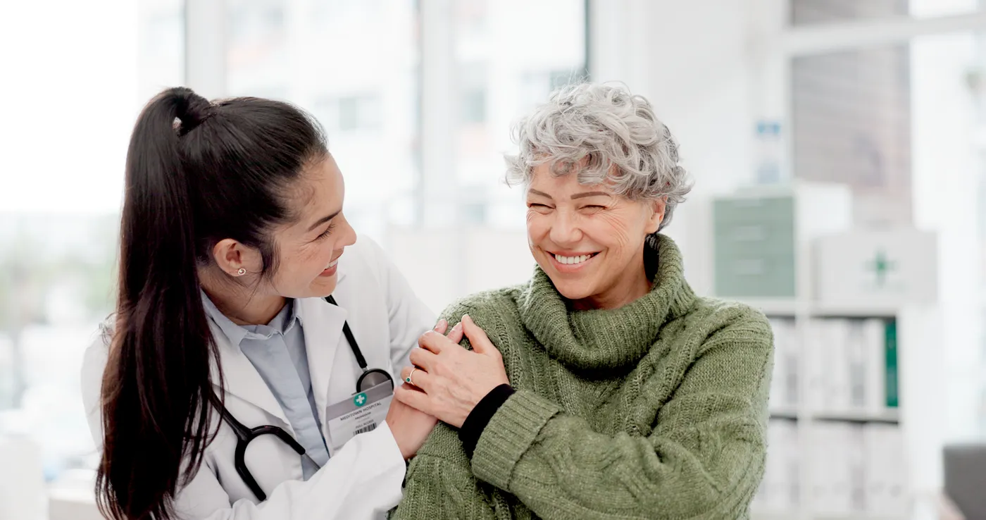 A clinician and an older patient sharing a laugh in a bright exam room.