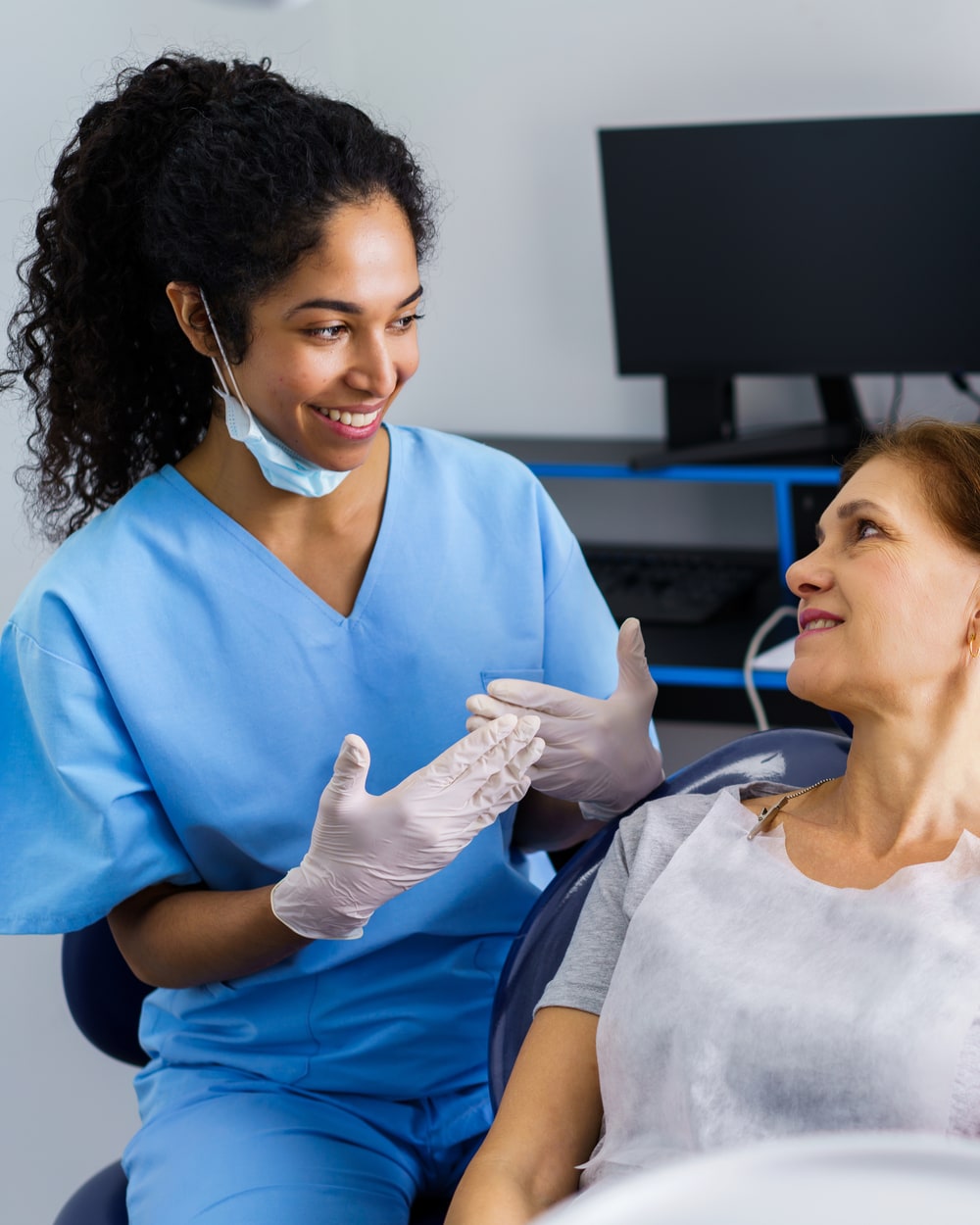A dental clinician and an older patient in unhurried conversation in a bright clinic — full attention, gloved hands mid-gesture, no screens between them.